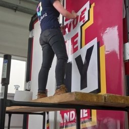 Copypoint - Pracownica na rusztowaniu aplikuje folię samoprzylepną z napisem na dużą powierzchnię reklamową w hali produkcyjnej.