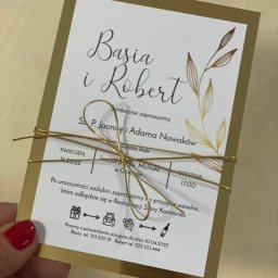 Copypoint - Eleganckie zaproszenie ślubne w odcieniach bieli i złota, przewiązane złotym sznurkiem, trzymane w dłoni z pomalowanymi na czerwono paznokciami.