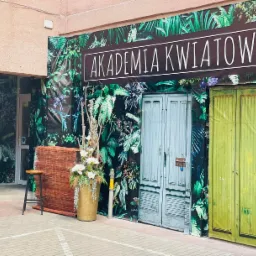Fasada budynku w Gnieźnie oklejona folią z motywem tropikalnych liści, na której umieszczono szyld 'Akademia Kwiatów'. Po lewej stronie wejście, a obok dekoracja z kwiatów w złotym wazonie.