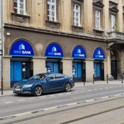 Elewacja budynku z niebieskimi szyldami Neo Bank, na pierwszym planie zaparkowany niebieski samochód Audi, w tle tramwajowe tory.
