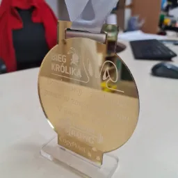 Złoty medal z grawerem 'Bieg Królika' na przezroczystej podstawce, zawieszony na szarej wstążce. Na medalu widoczne logo biegu oraz podziękowanie za pomoc w organizacji. W tle biurko z klawiaturą...