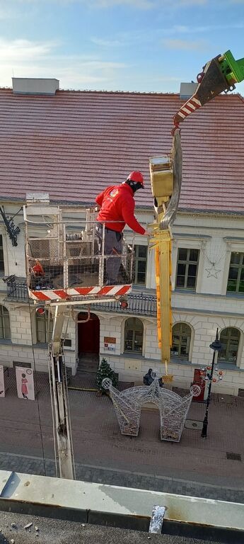 Pracownik w czerwonej kurtce i kasku montuje dekoracje świąteczne na ulicznej latarni, używając podnośnika koszowego; w tle budynek z czerwoną dachówką i ozdobna brama świetlna w kształcie korony.