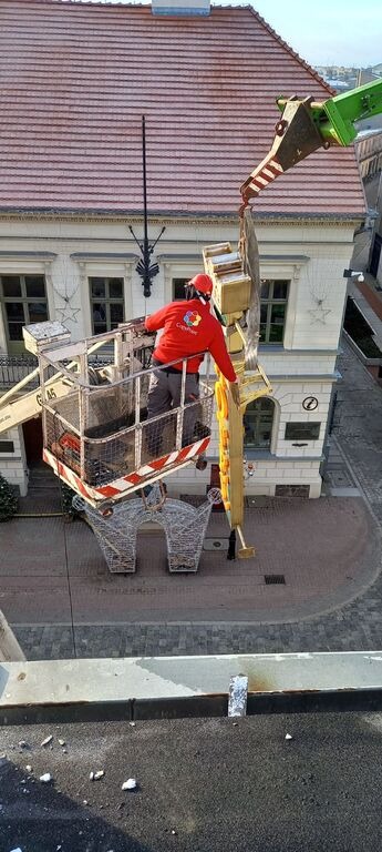 Montaż świątecznych dekoracji ulicznych za pomocą dźwigu koszowego, pracownik w czerwonej kurtce i kasku, ozdoby w kolorze złotym, budynek w tle.