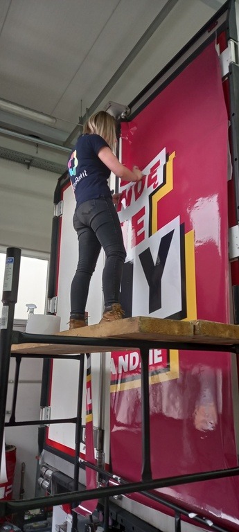 Pracownica na rusztowaniu aplikuje folię samoprzylepną z napisem na dużą powierzchnię reklamową w hali produkcyjnej.