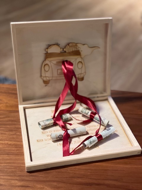 Drewniane pudełko na prezent ślubny z wygrawerowanym samochodem i młodą parą, banknoty zwinięte i przewiązane bordową wstążką, dedykacja wygrawerowana w drewnie.