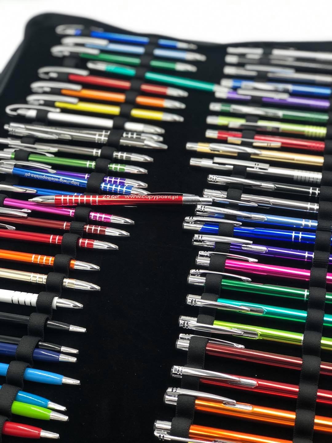 Kolekcja kolorowych długopisów z metalowymi elementami, umieszczonych w czarnym etui z elastycznymi uchwytami, jeden długopis z nadrukowanym adresem strony internetowej.
