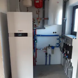 Biały piec grzewczy Viessmann z podłączonymi instalacjami, w tym czerwony zbiornik wyrównawczy i biały zasobnik ciepłej wody, widoczne rury miedziane i plastikowe, filtry i zawory, zamontowane...
