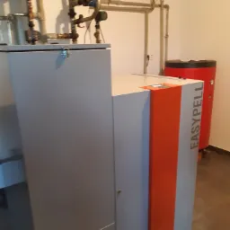 Szary piec z pomarańczowym akcentem marki Easypell w kotłowni z widocznymi rurami i czerwonym zasobnikiem ciepłej wody.