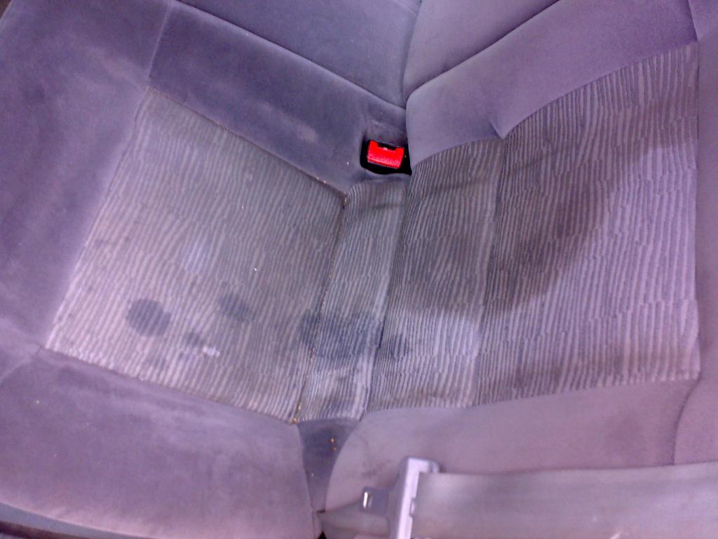 Zabrudzone tylne siedzenie samochodu z plamami przed praniem tapicerki, widoczny fragment pasa bezpieczeństwa i czerwony zatrzask.