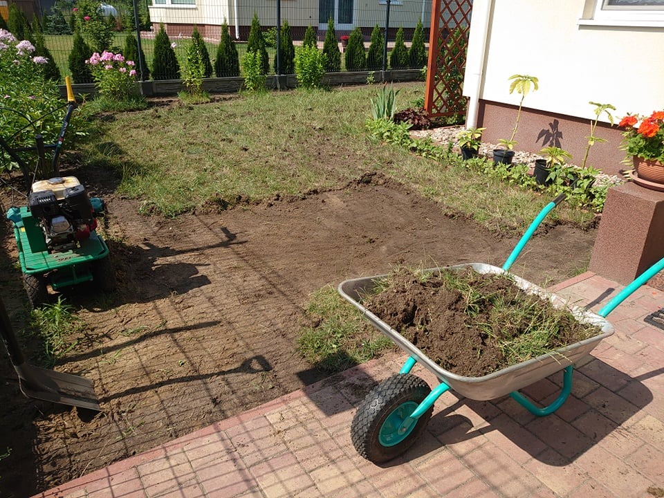 Przygotowanie terenu pod trawnik: glebogryzarka, taczka z ziemią i szpadel na tle częściowo wyrównanego terenu z fragmentami trawy.