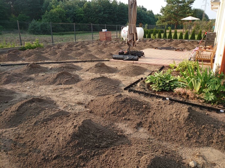 Przygotowanie terenu pod ogród w Legionowie: widok na nierówny teren z kopcami ziemi, czarne obrzeża rabat, w tle siatka ogrodzeniowa i drzewa.