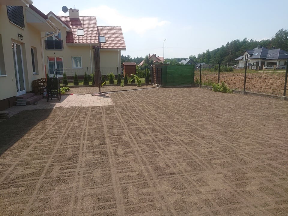 Przygotowany teren pod trawnik z widocznymi śladami po walcowaniu, obok dom z czerwonym dachem i młode tuje wzdłuż ogrodzenia.