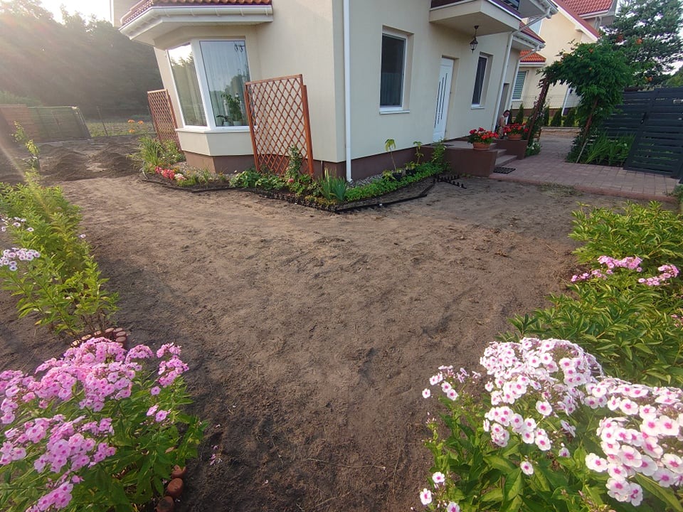 Przygotowany teren pod nowy ogród z widocznymi rabatami kwiatowymi, siatkami podporowymi i świeżo posadzoną roślinnością przy domu jednorodzinnym w słoneczny dzień.