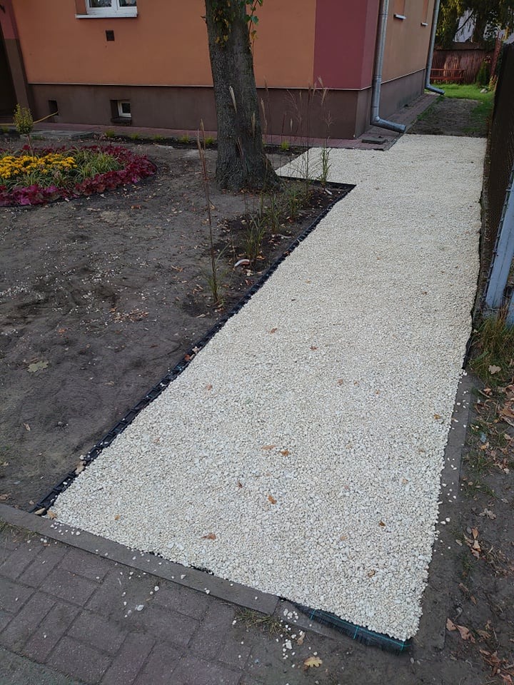 Ścieżka z jasnego żwiru, wykończona czarną obwódką, prowadząca przez ogród z rabatą kwiatową i młodymi drzewkami, widok z góry.