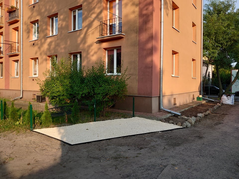 Przygotowany teren pod trawnik lub ogródek przed budynkiem mieszkalnym, wyłożony białym żwirem, otoczony czarną obwódką, z widoczną roślinnością w tle i fragmentem elewacji budynku.