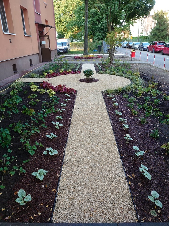 Kształtny chodnik z jasnego żwiru prowadzący przez rabaty z korą i roślinami ozdobnymi, widok na budynek i ulicę.