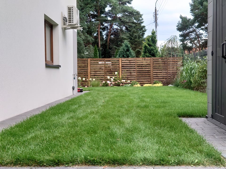 Starannie utrzymany trawnik przylegający do białego budynku z drewnianą ramą okienną i klimatyzatorem na ścianie, z drewnianym płotem i zielenią w tle, widok z perspektywy chodnika.