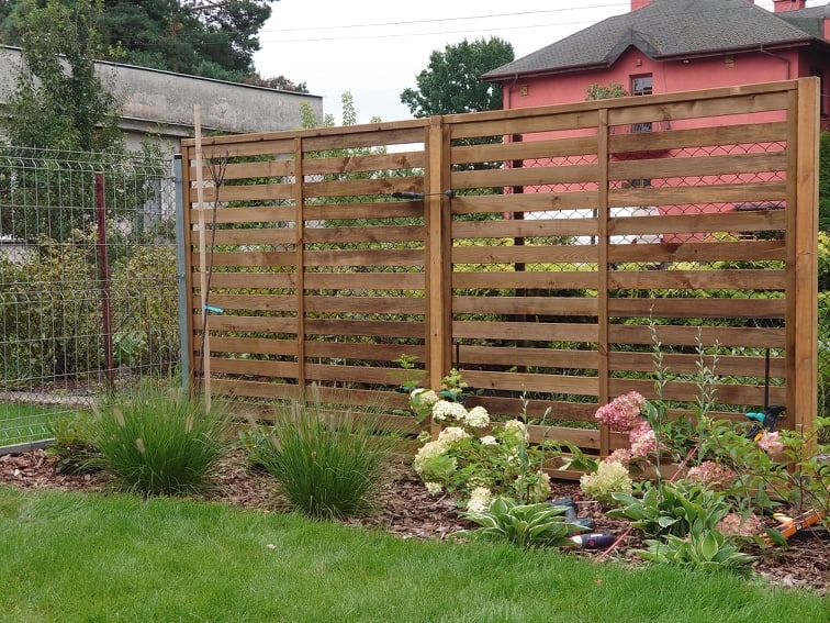 Drewniany ażurowy płot oddzielający ogród z kwitnącymi hortensjami i ozdobnymi trawami od posesji sąsiada z siatką ogrodzeniową.