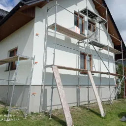 Dom jednorodzinny w trakcie prac elewacyjnych, widoczne rusztowanie i biała, świeżo pomalowana fasada, z pomarańczowymi zaślepkami, w tle błękitny basen.