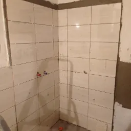 Ściana narożna w trakcie układania jasnych płytek ceramicznych, widoczne krzyżyki dystansowe i wystające zawory do baterii.