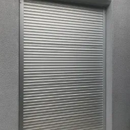 Zamknięta, srebrna roleta zewnętrzna na tle szarej, chropowatej ściany budynku, widoczny fragment parapetu wyłożonego płytkami.