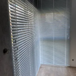 Jasne, drewnopodobne żaluzje poziome zamontowane w oknach, widoczne sznurki sterujące po lewej stronie, podłoga z paneli, surowa ściana z wystającą instalacją elektryczną.