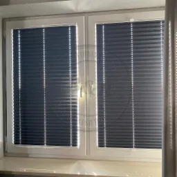 Zamknięte, grafitowe plisy okienne w białej ramie okiennej, widoczne z bliska, z delikatnym światłem słonecznym przebijającym przez lamele.