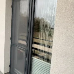 Zamknięta moskitiera ramkowa w kolorze antracytowym na drzwiach balkonowych, obok roleta plisowana, widok z zewnątrz na szorstką elewację budynku.