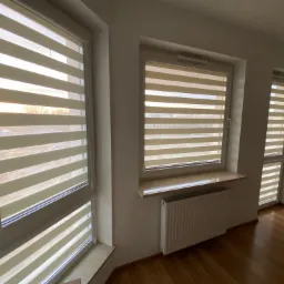 Trzy okna z zamontowanymi roletami w systemie dzień i noc, widoczne poziome pasy materiału, jasne wnętrze z drewnianą podłogą i grzejnikiem pod jednym z okien.