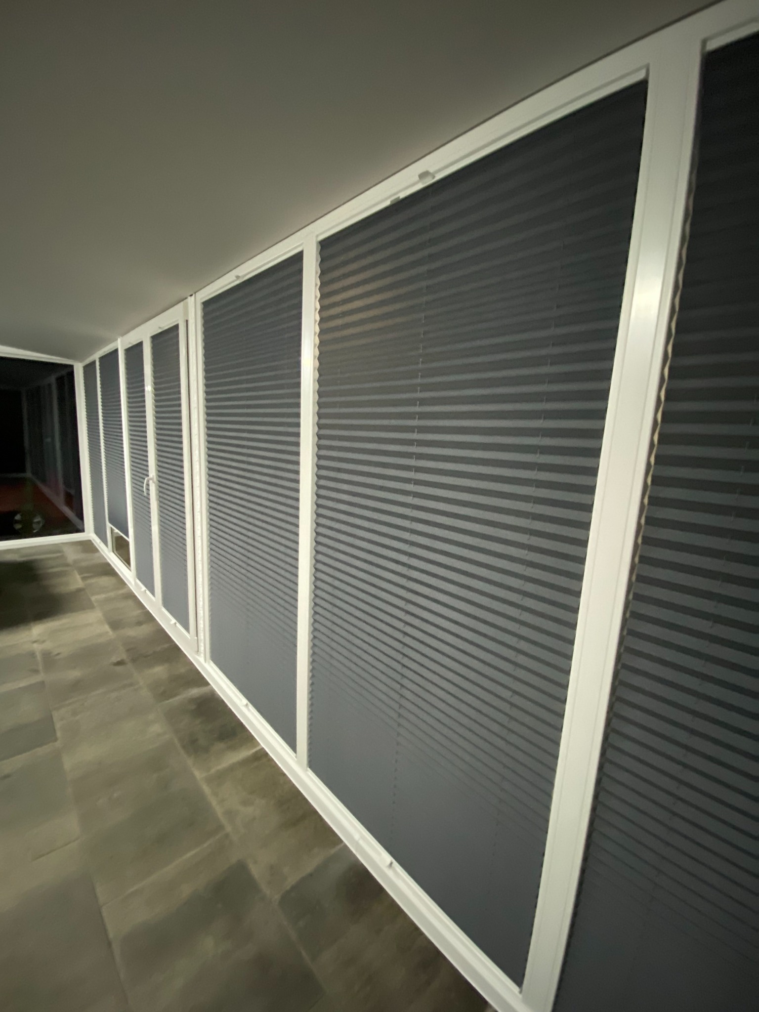 Szare, plisowane osłony okienne z białymi ramami, zamontowane wzdłuż ściany z oknami, widok z boku, z widoczną podłogą z kamiennych płyt.