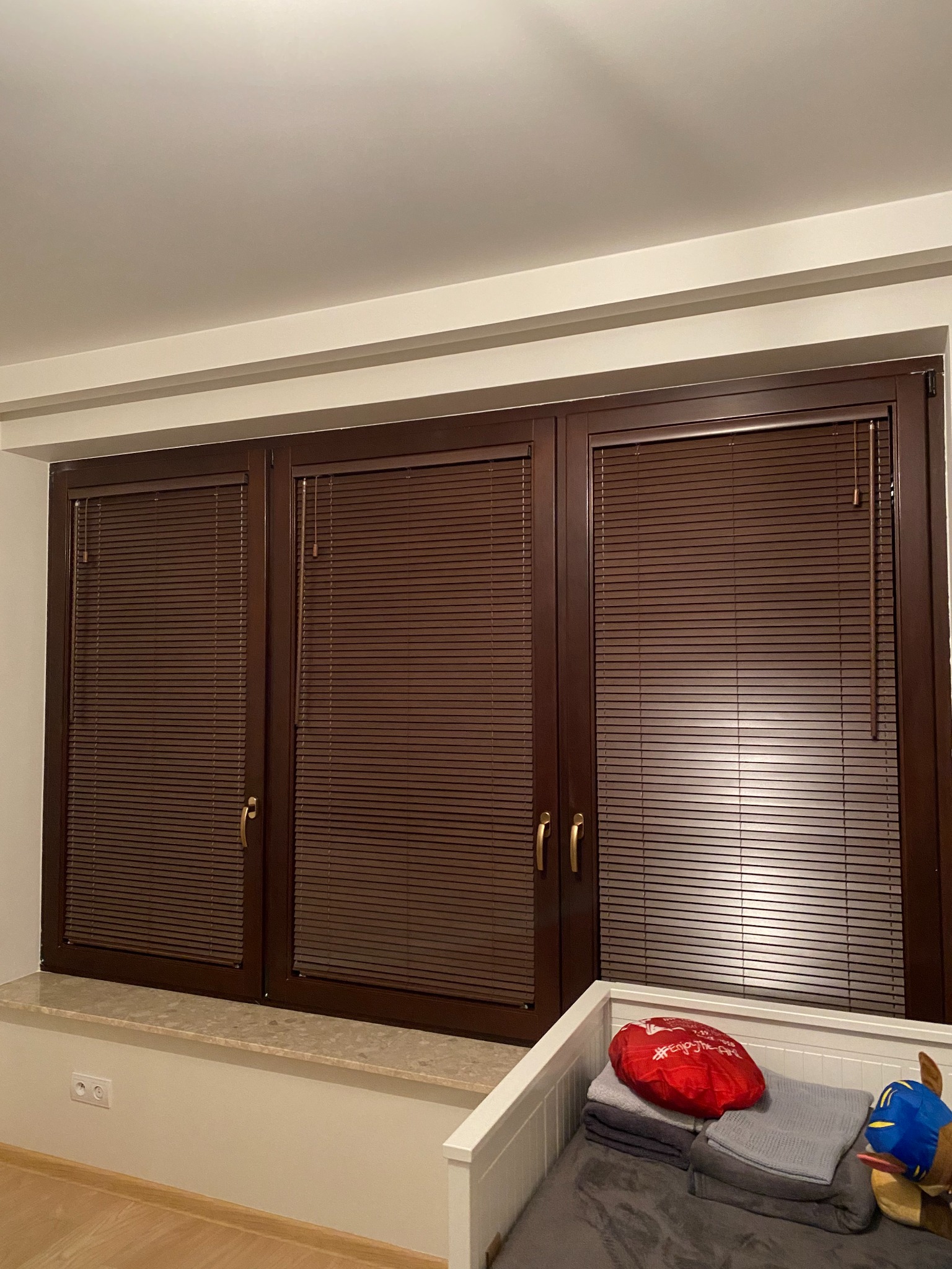 Trzy okna z brązowymi, poziomymi listwami w pokoju dziecięcym, widoczne łóżeczko z pościelą i zabawką.