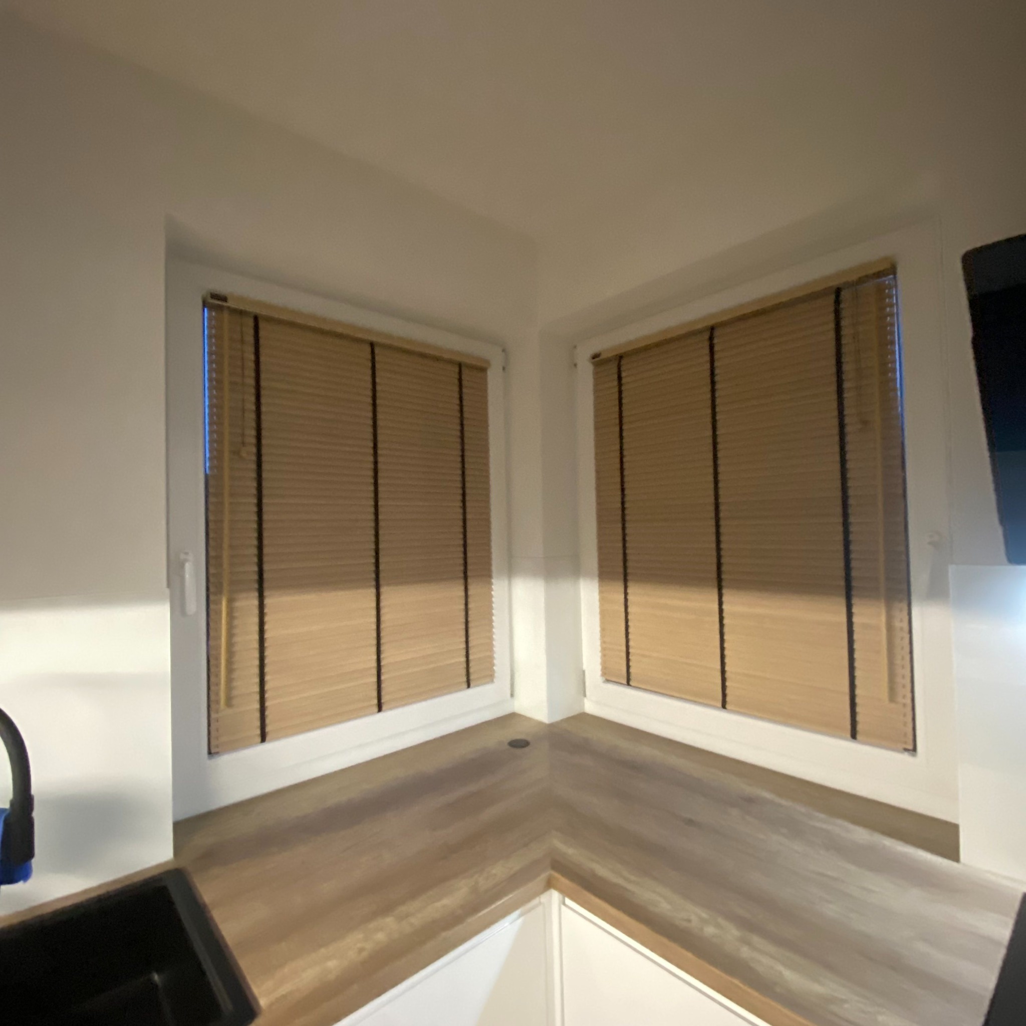 Dwa okna narożne z zamontowanymi plisami w kolorze jasnego drewna, widoczne z perspektywy blatu kuchennego.