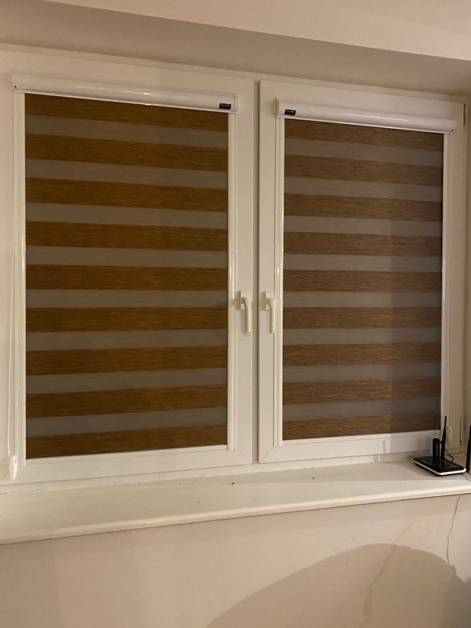 Dwa okna z zamontowanymi roletami w systemie dzień i noc, widoczne poziome pasy w odcieniach brązu i beżu, białe ramy okienne, na parapecie leżący telefon i router.