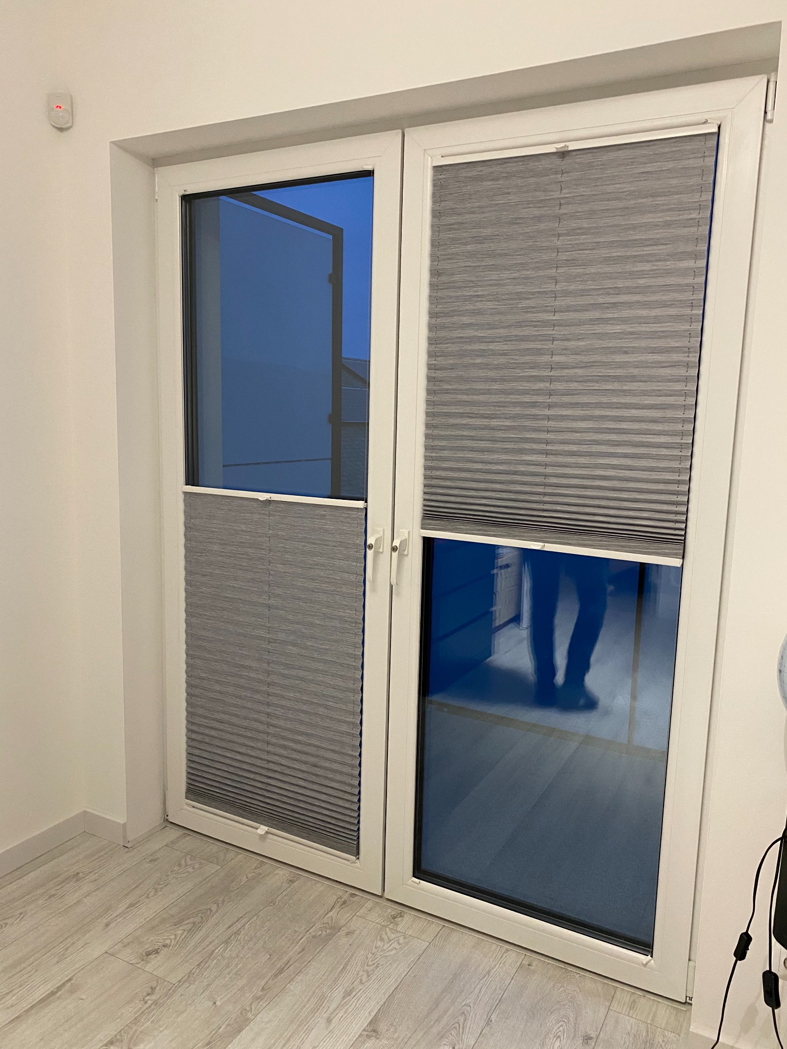 Białe drzwi balkonowe z szarymi plisami częściowo zasłoniętymi, widoczne odbicie osoby w szybie.