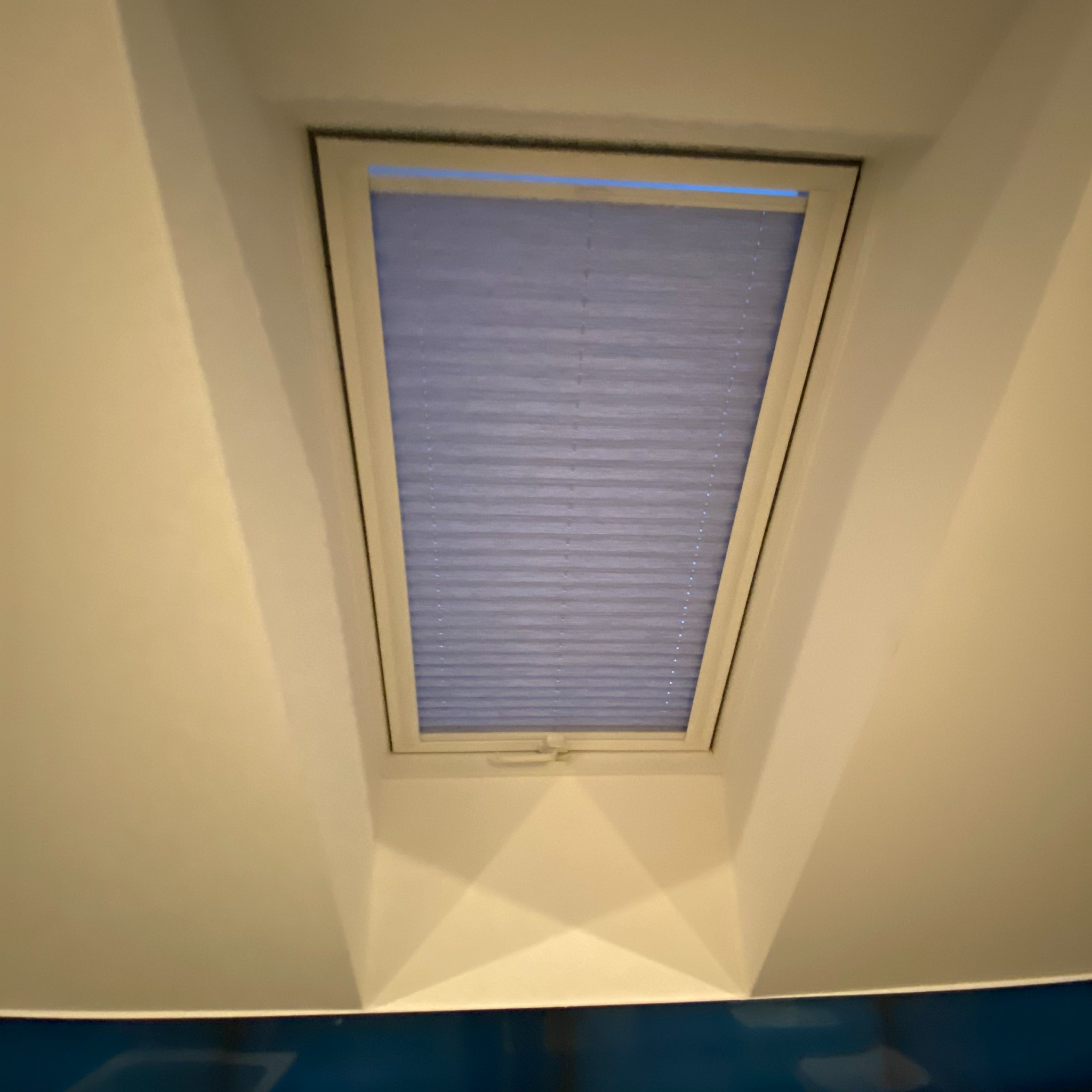 Zamknięta, jasnoniebieska przesłona okienna typu plisa w oknie dachowym, widok od dołu, białe ramy okienne i ściany.