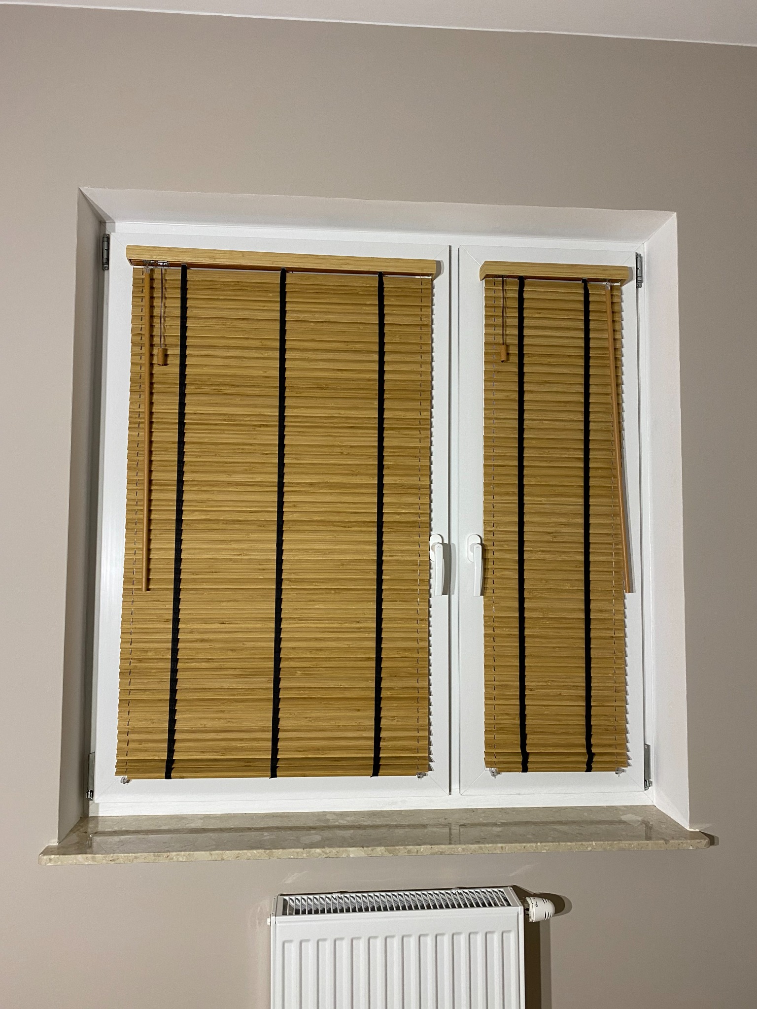 Bambusowe lamele w oknie PCV, z czarnymi sznurkami, częściowo opuszczone, widoczny biały grzejnik pod parapetem.