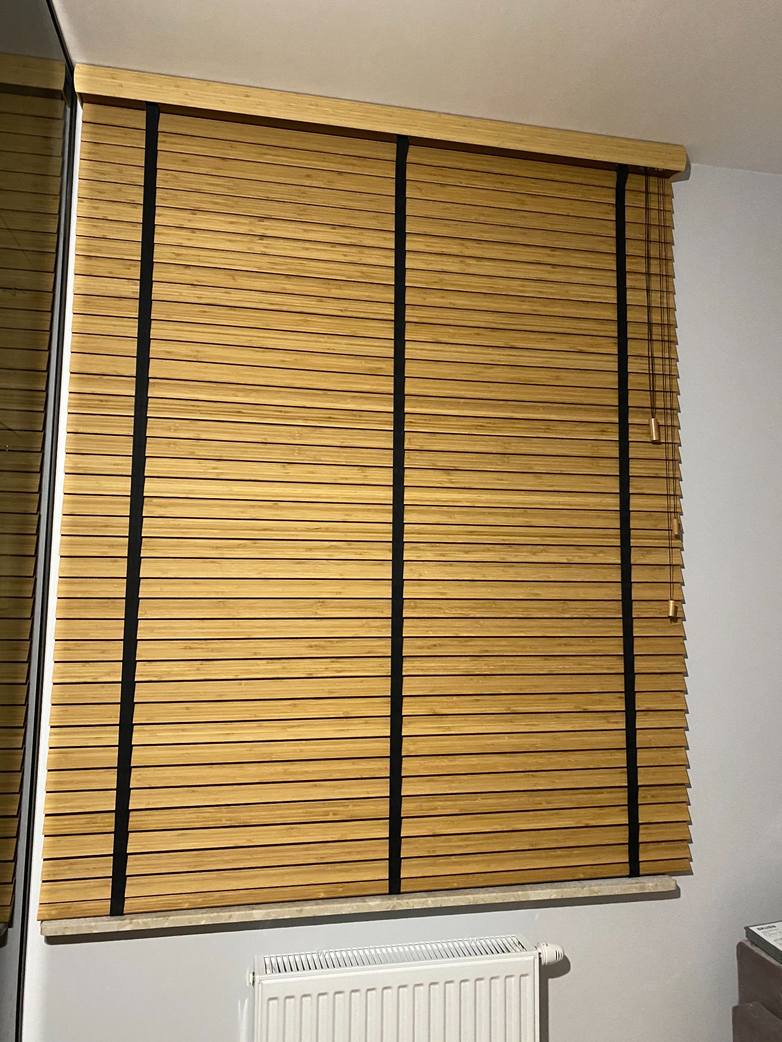 Zamknięte drewniane żaluzje w odcieniach brązu z czarnymi paskami mocującymi, zamontowane w oknie, widoczny fragment grzejnika pod parapetem.
