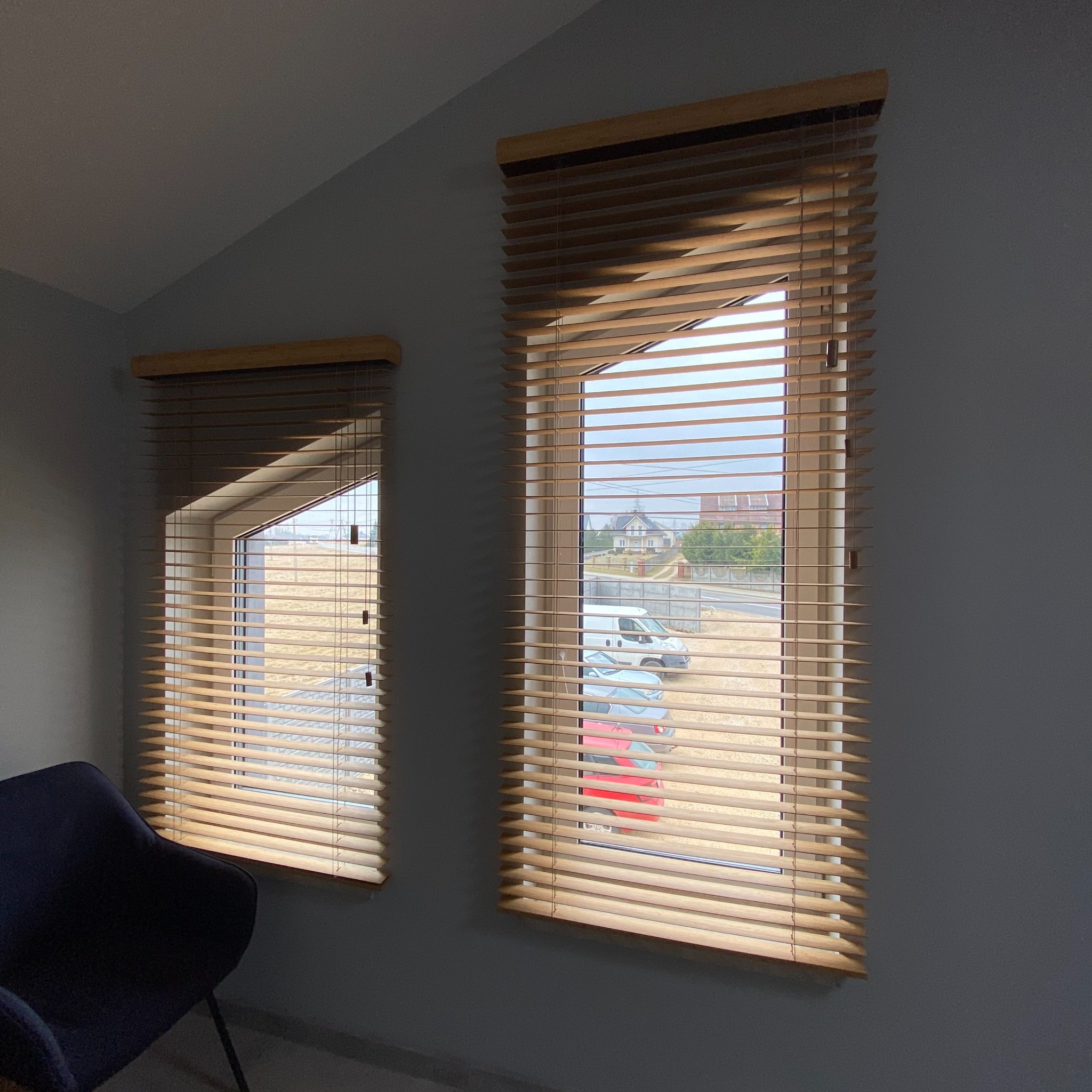 Dwa okna z drewnianymi listwami żaluzji w odcieniach beżu, z widokiem na dom i samochody na zewnątrz, na tle szarej ściany.