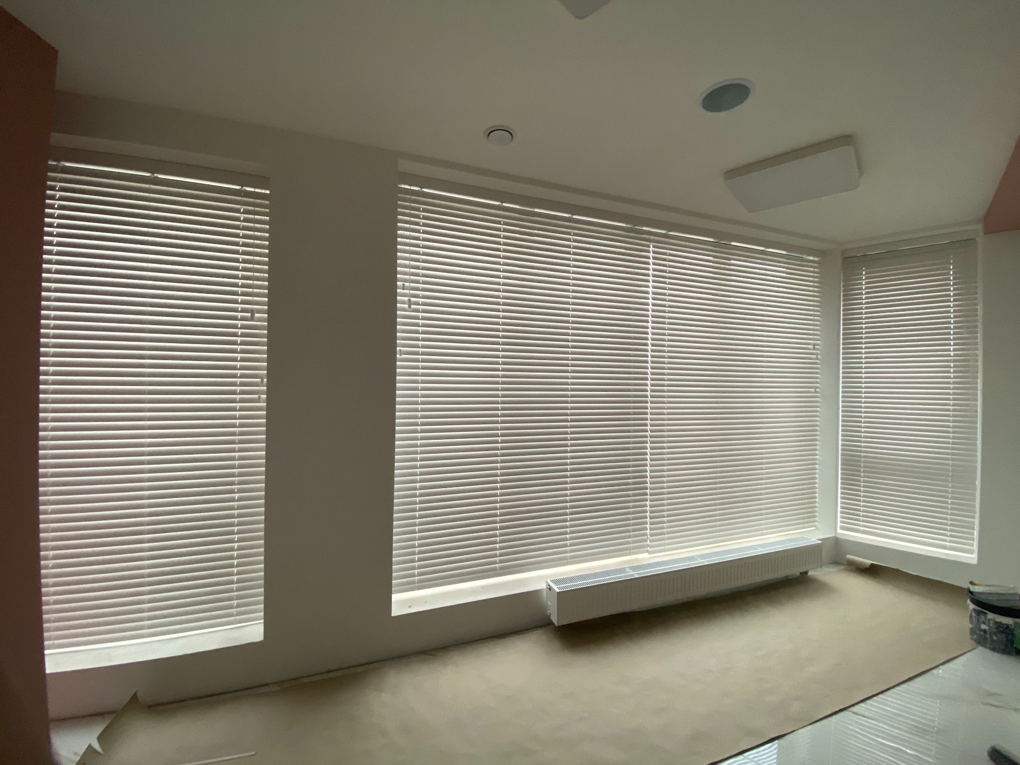 Wnętrze pokoju z trzema oknami wyposażonymi w poziome, jasne listewki, prawdopodobnie osłony okienne, z widocznym grzejnikiem pod jednym z okien i zabezpieczoną podłogą.