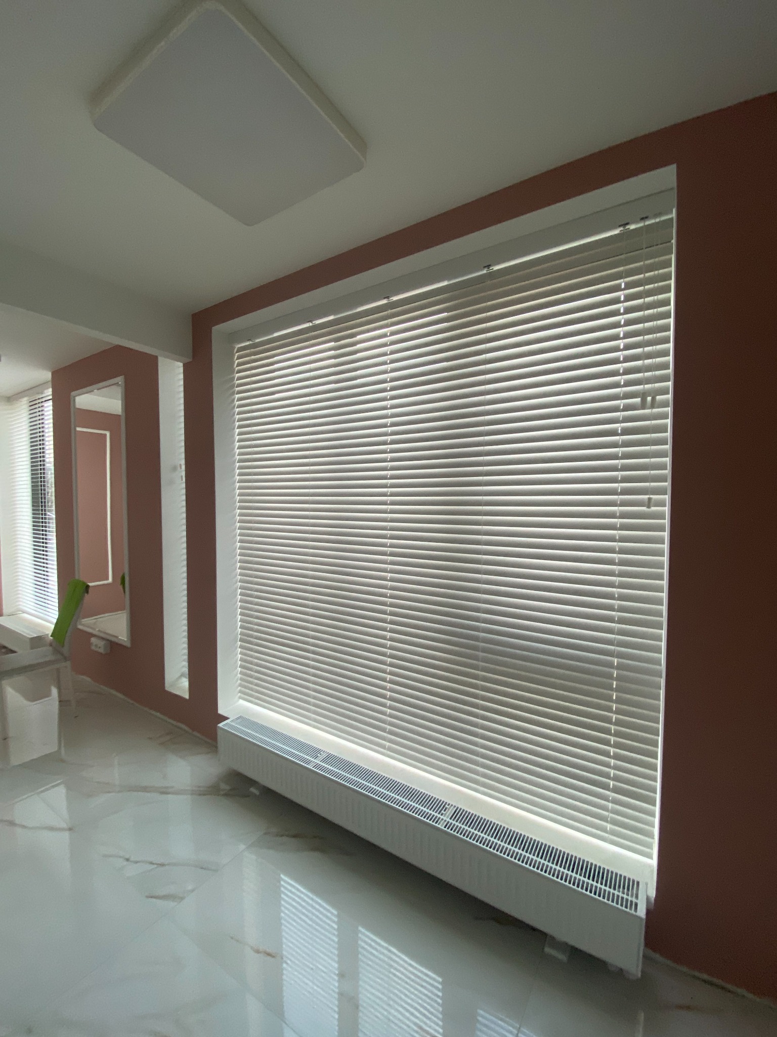 Wnętrze pokoju z białymi, poziomymi przesłonami okiennymi, częściowo zasłoniętymi, zamontowanymi nad grzejnikiem. Widoczne odbicia w polerowanej podłodze i fragment lustra w tle.