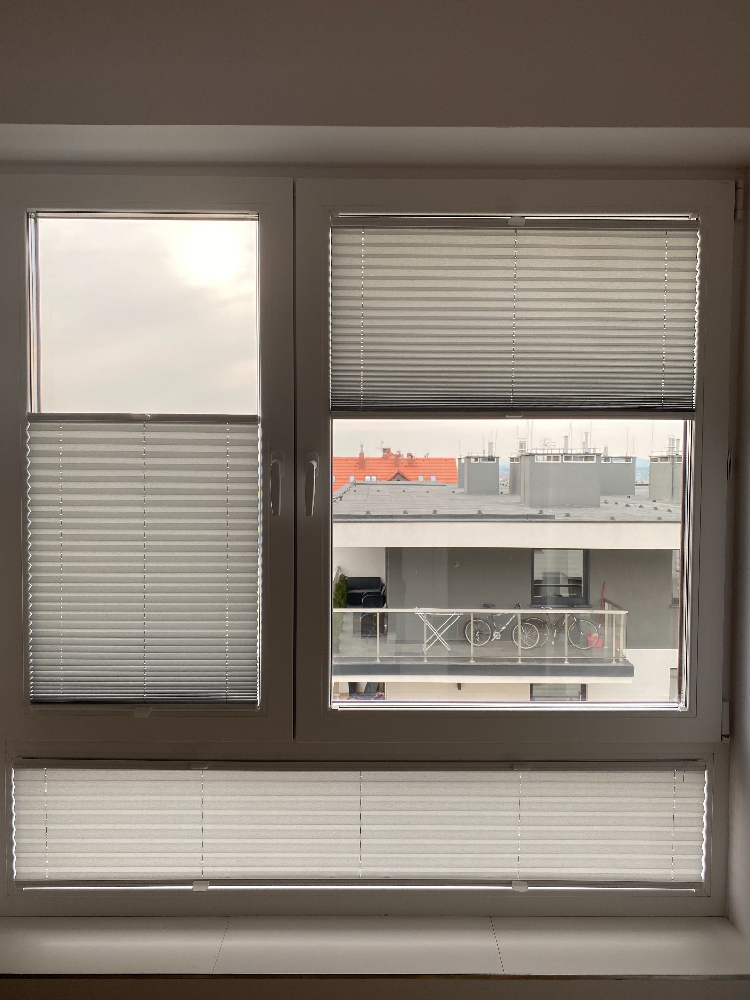 Białe okno z trzema segmentami przesłoniętymi plisowanymi roletami w różnym stopniu, widok na budynek mieszkalny z balkonem i rowerami.