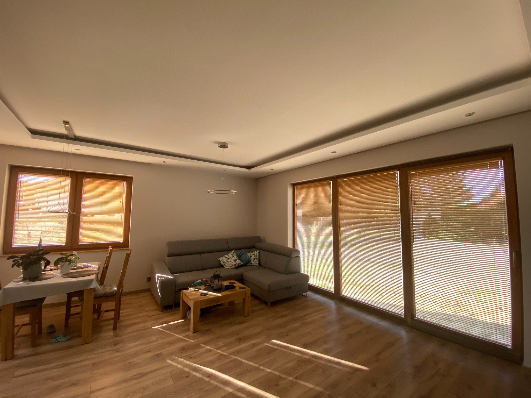 Jasne wnętrze salonu z drewnianą podłogą, szarą sofą narożną i oknami przesłoniętymi poziomymi osłonami okiennymi w kolorze drewna, wpadające promienie słoneczne tworzą grę światła i cienia.