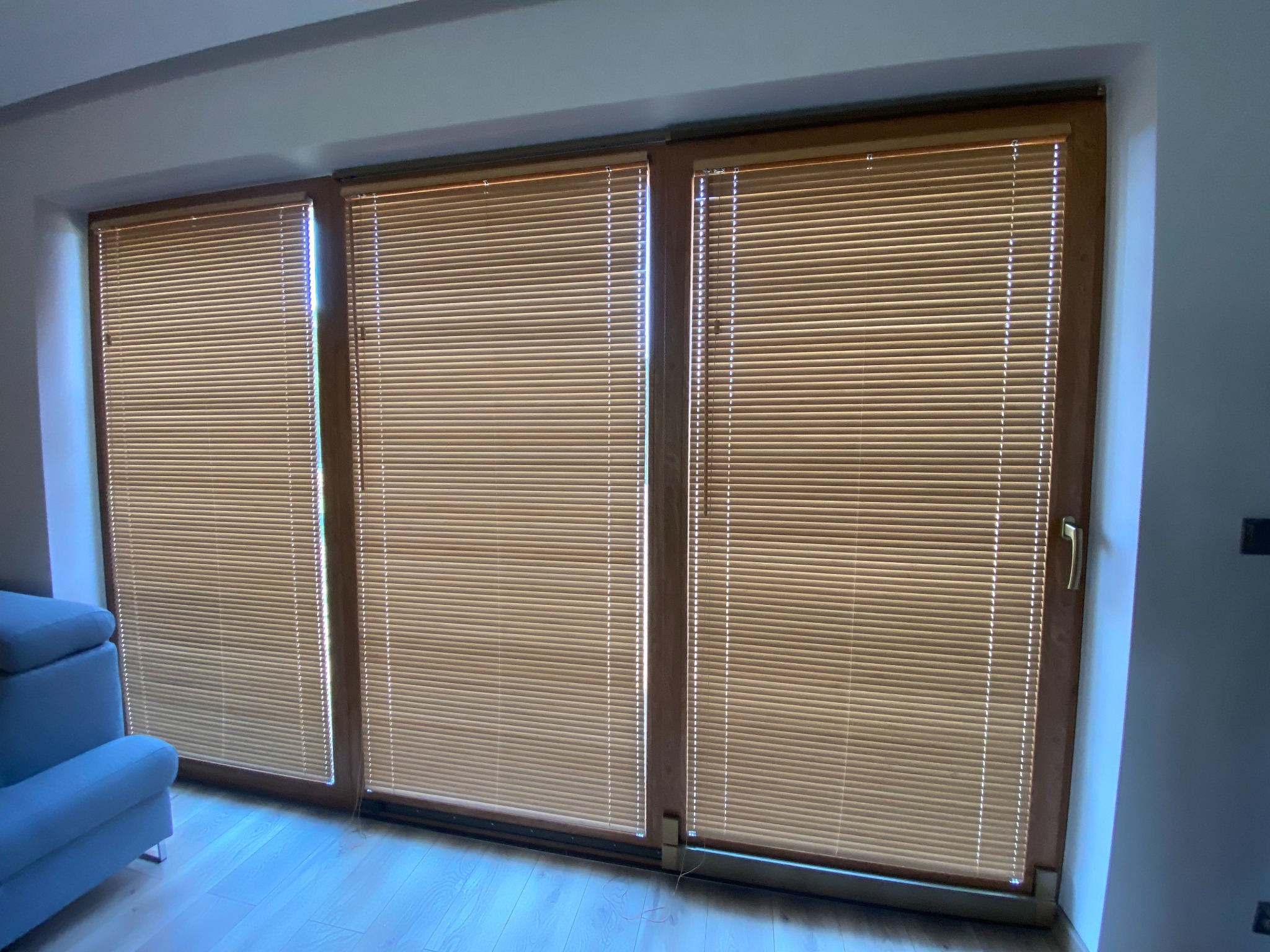 Trzy okna z drewnianymi ramami, zasłonięte poziomymi żaluzjami w odcieniach beżu, widok z wnętrza pomieszczenia z fragmentem niebieskiej sofy.