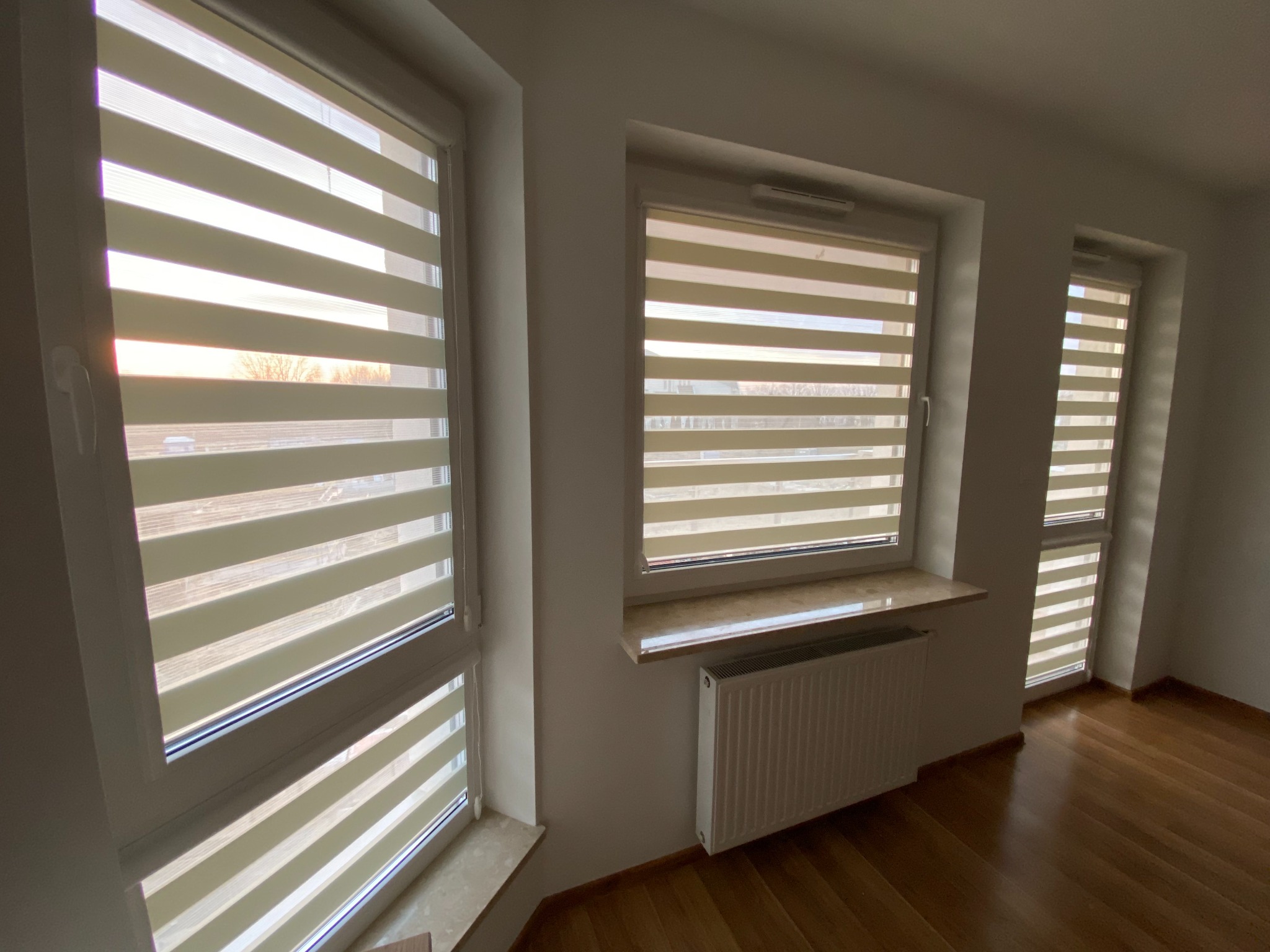Trzy okna z zamontowanymi roletami w systemie dzień i noc, widoczne poziome pasy materiału, jasne wnętrze z drewnianą podłogą i grzejnikiem pod jednym z okien.