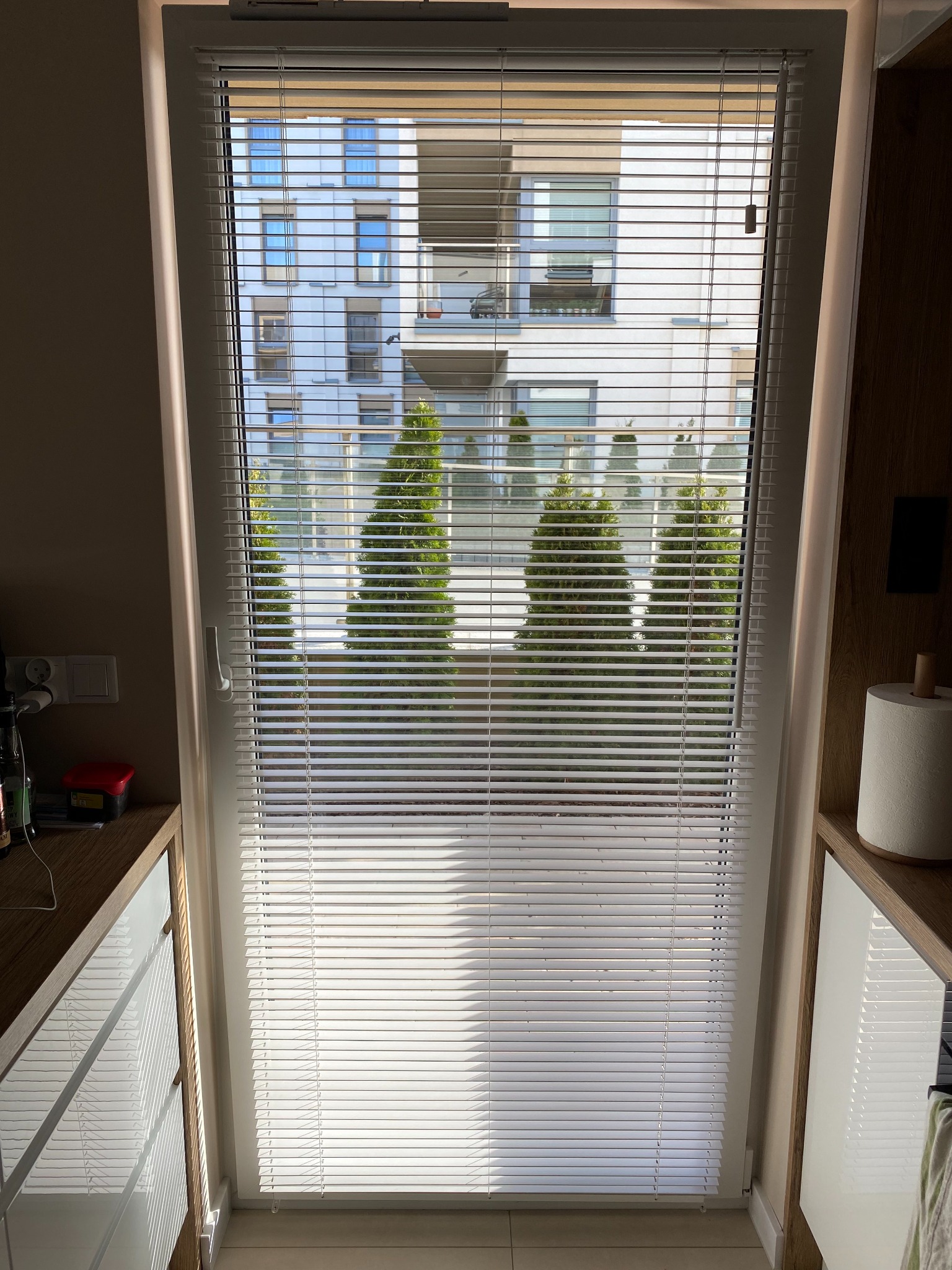 Zamknięte białe żaluzje aluminiowe w oknie, widok na nowoczesny budynek i drzewa iglaste na zewnątrz.