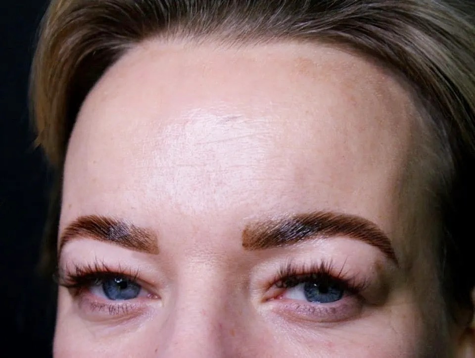 Brwi po zabiegu laminacji, widoczne sztuczne rzęsy i niebieskie oczy, ciemne tło.