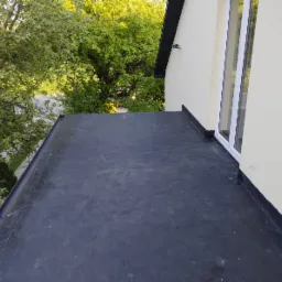 Widok z góry na płaski dach pokryty czarną membraną bitumiczną, z widoczną ścianą budynku w kremowym kolorze i oknem PCV, oraz bujną zielenią drzew w tle.