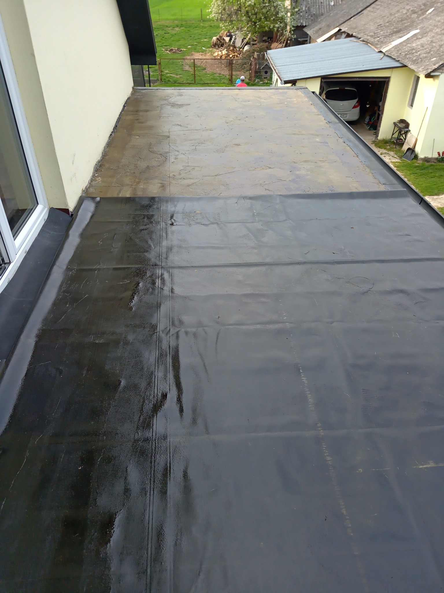 Płaski dach pokryty czarną membraną bitumiczną, widoczne łączenia i fragment starego pokrycia, w tle budynek gospodarczy z garażem.