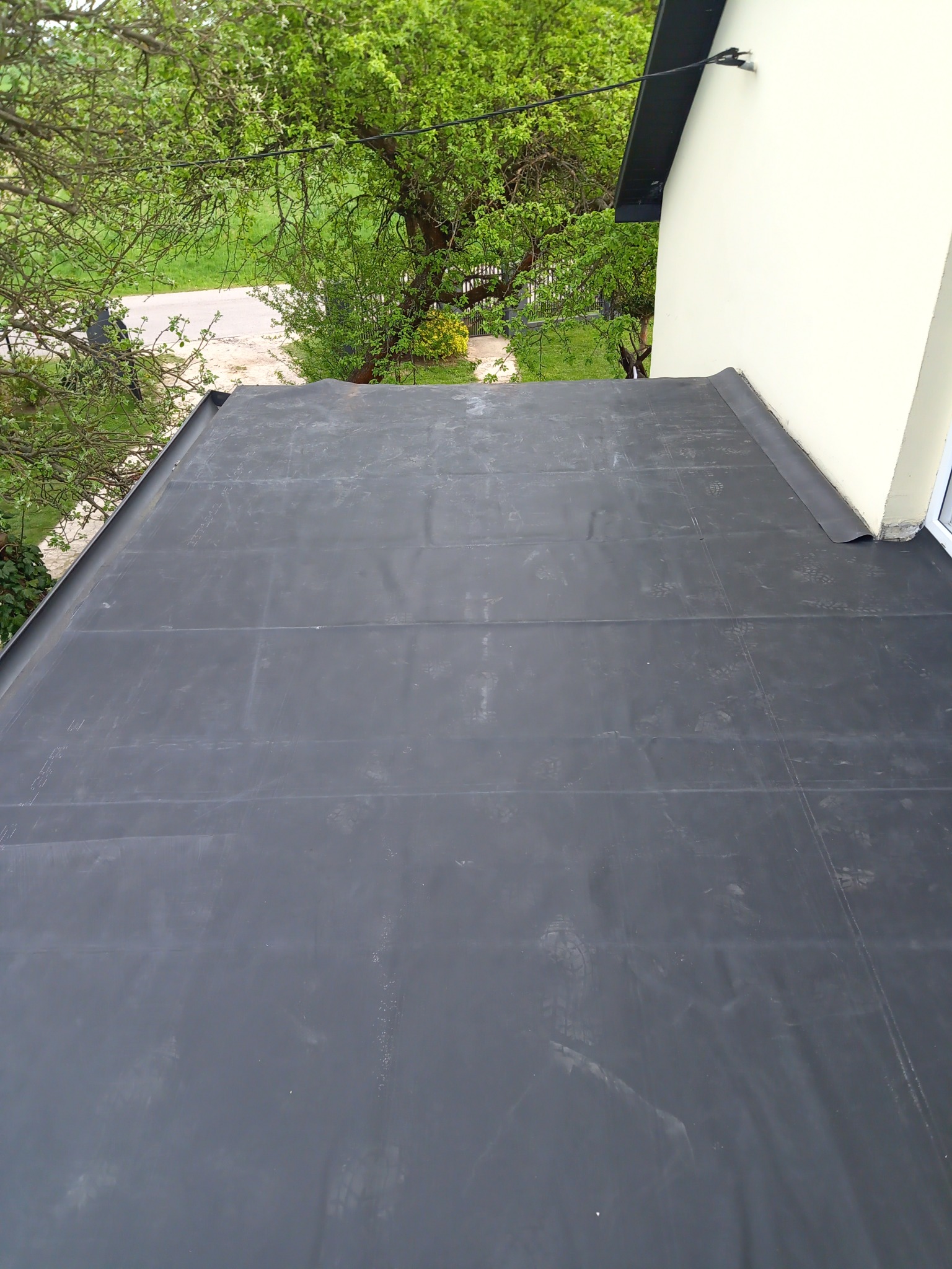 Widok z góry na płaski dach pokryty czarną papą termozgrzewalną, widoczne łączenia papy i fragment ściany budynku z oknem oraz drzewa w tle.