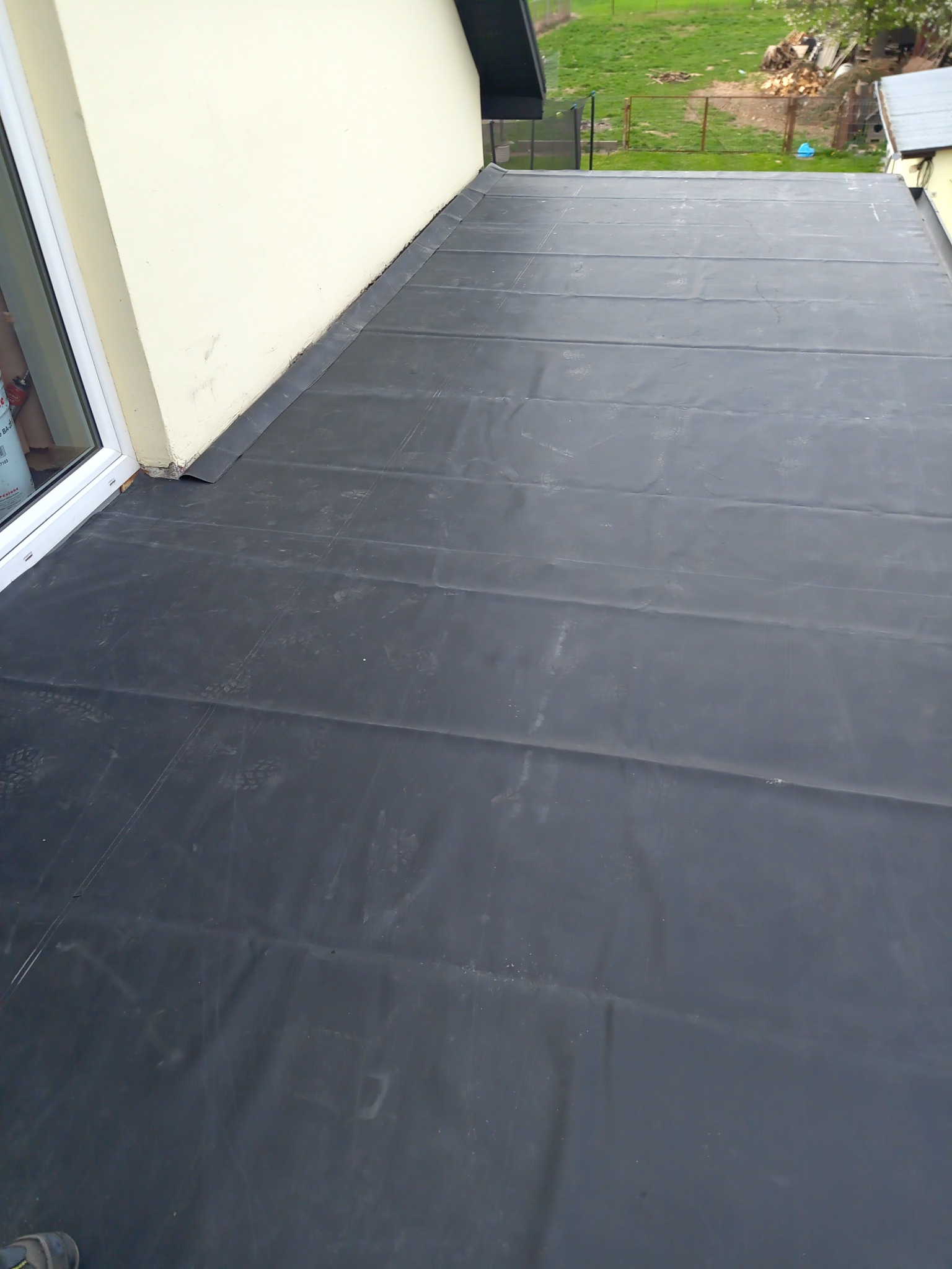 Czarna papa termozgrzewalna na płaskim dachu budynku w Wieniawie, widok z góry, z fragmentem okna i zielonym otoczeniem w tle.